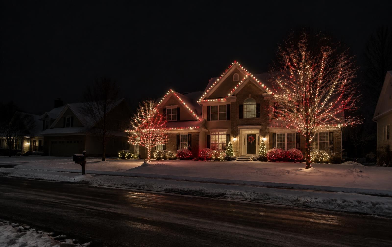 Colonial home lit in red and warm white lights with wrapped trees on a snowy street